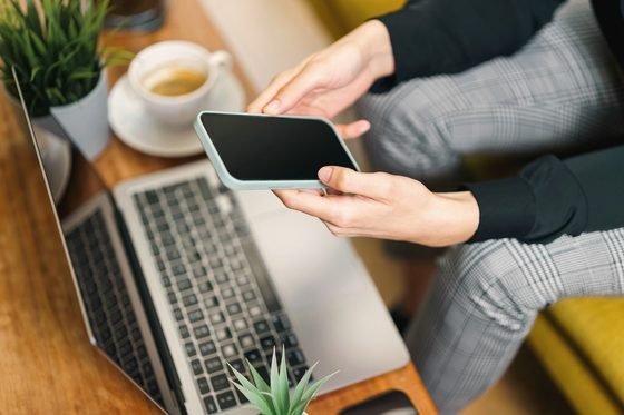 Dos manos con un celular de pantalla en blanco sobre una laptop abierta, junto a una taza de café.